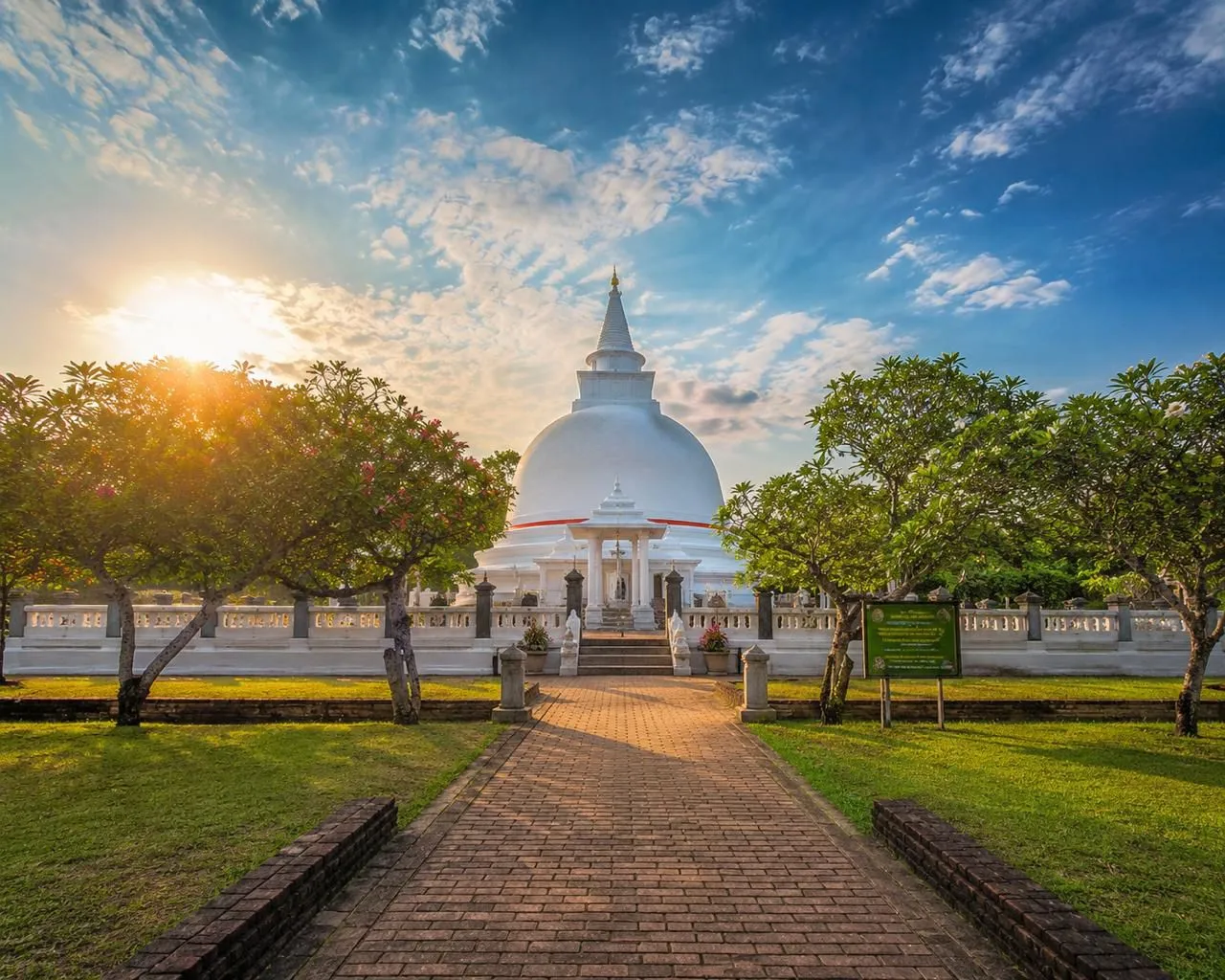 A brick path lined with frangipani trees leading to a white domed stupa