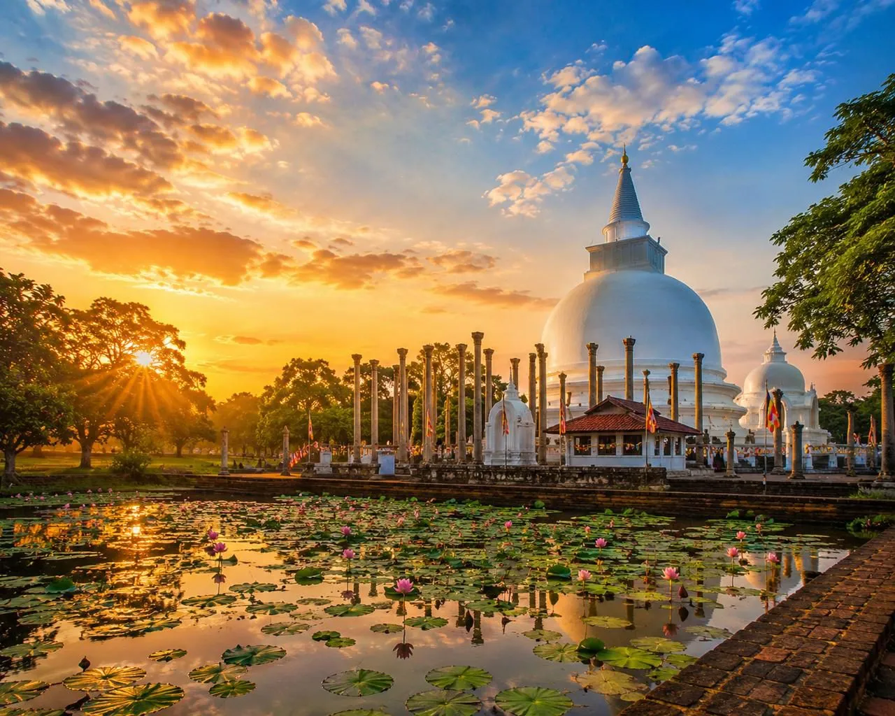 A white stupa ringed by ancient stone pillars, reflected in a lotus pond at sunset