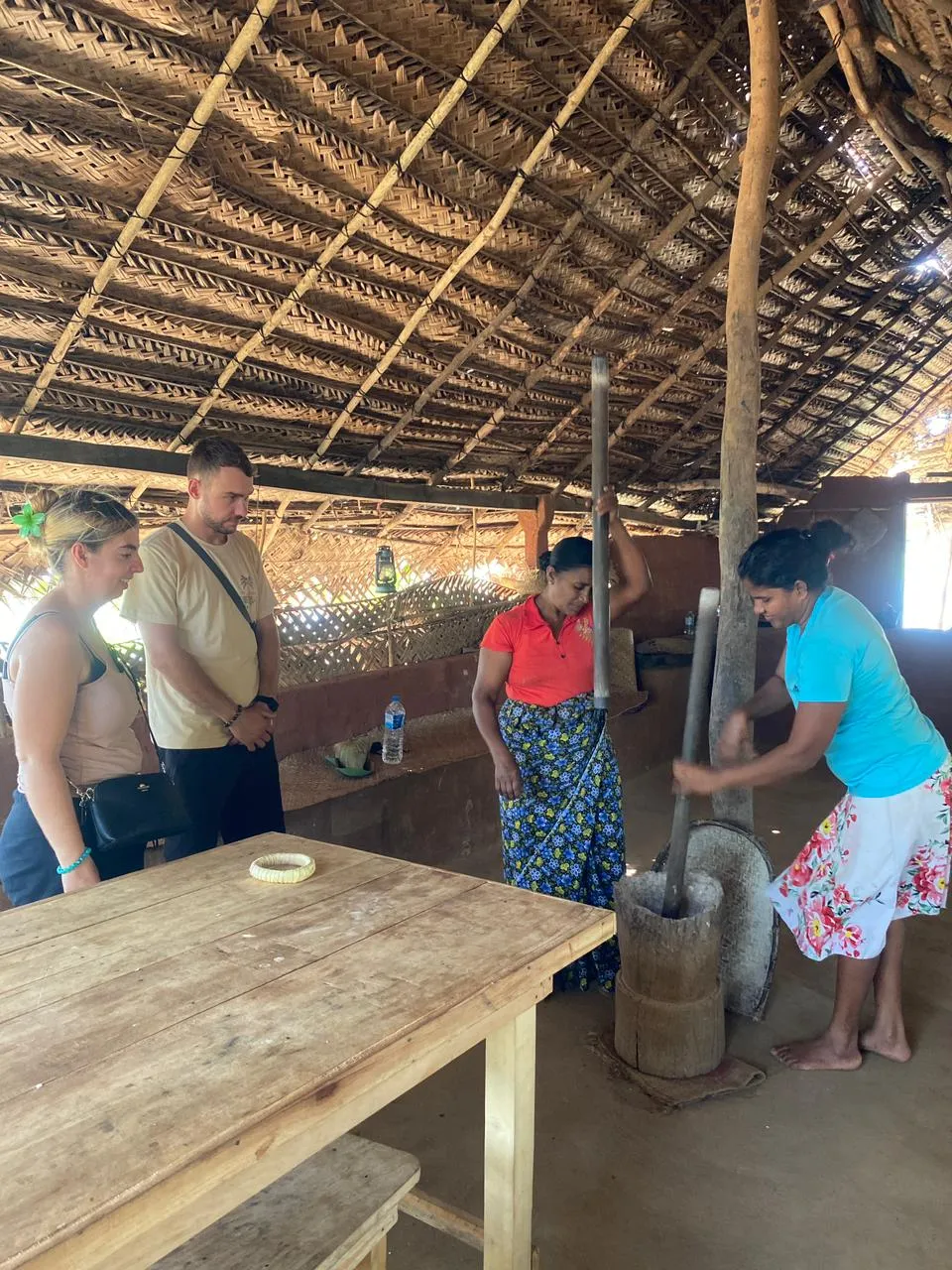 Village women pounding rice with wooden pestles while guests watch under a thatched roof